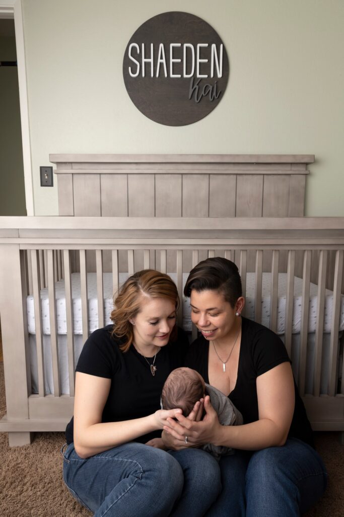 same sex female couple holding newborn boy while sitting in front of his crib