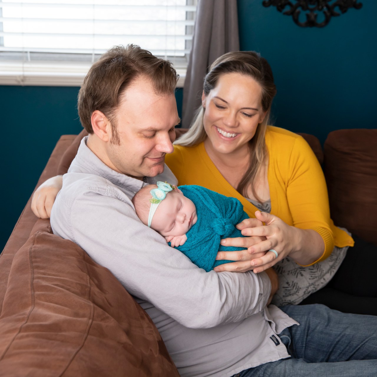 parents sitting on couch and holding newborn baby, mom wearing bright yellow, baby in teal, dad in lavender blue